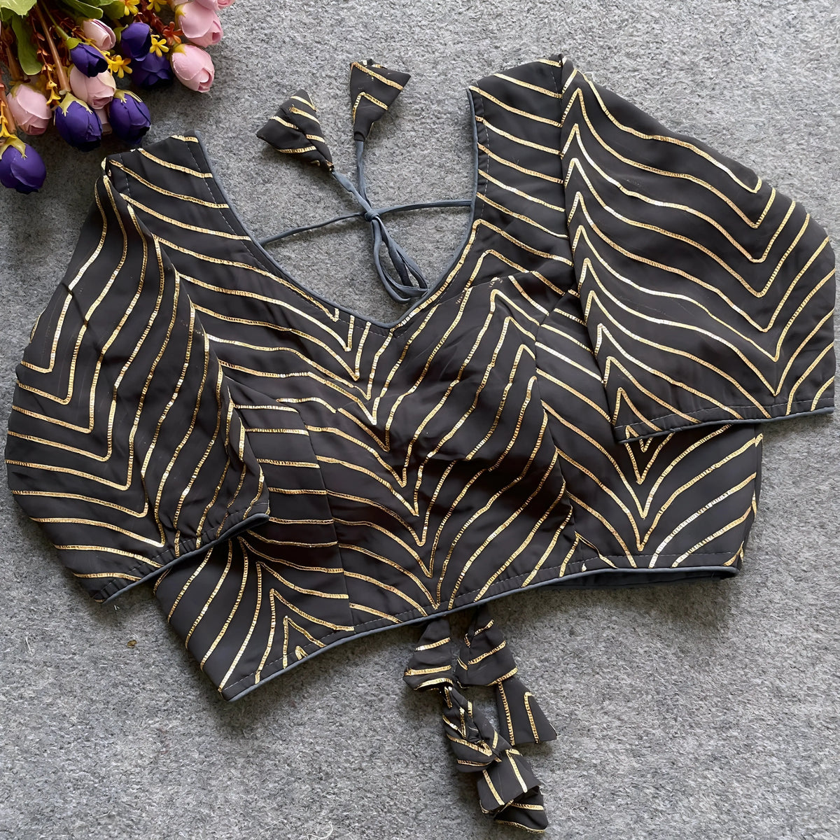 Black blouse featuring striking gold gota patti zigzag embroidery, three-quarter sleeves, and a stylish back tie-up with coordinated triangular tassels. Displayed flat on a grey textured surface alongside pink and purple flowers, emphasizing its festive and elegant ethnic appearance.