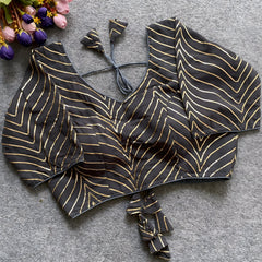 Black blouse featuring striking gold gota patti zigzag embroidery, three-quarter sleeves, and a stylish back tie-up with coordinated triangular tassels. Displayed flat on a grey textured surface alongside pink and purple flowers, emphasizing its festive and elegant ethnic appearance.
