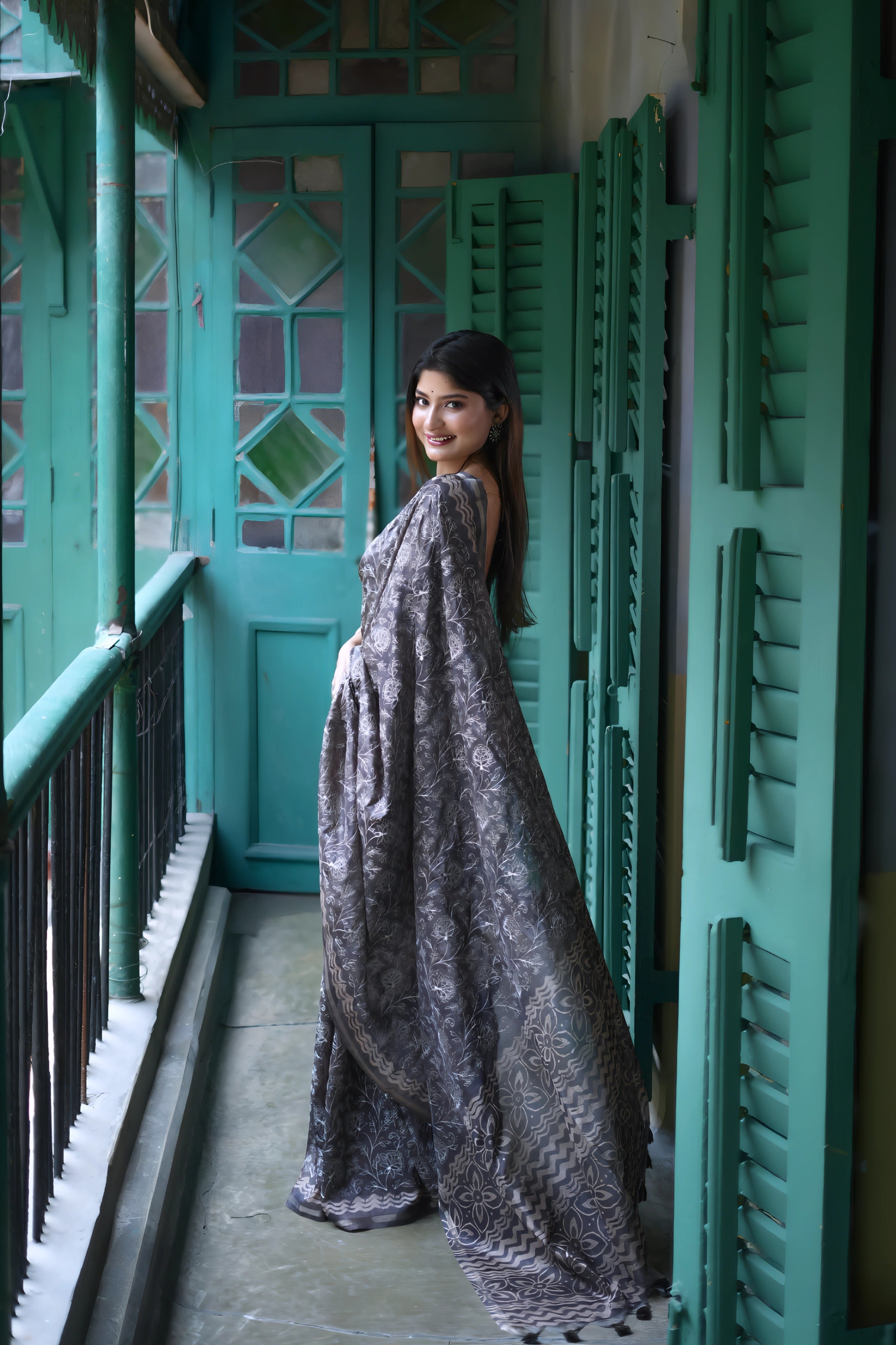 A woman models an elegant Black Tussar Silk Saree. The saree features dense all-over floral embroidery/weaving in a shimmering metallic silver thread. She wears a black sheer-sleeve blouse and heavy oxidized silver jewelry, including a large ring and stacked bangles, posing in a doorway framed by vibrant teal shutters.