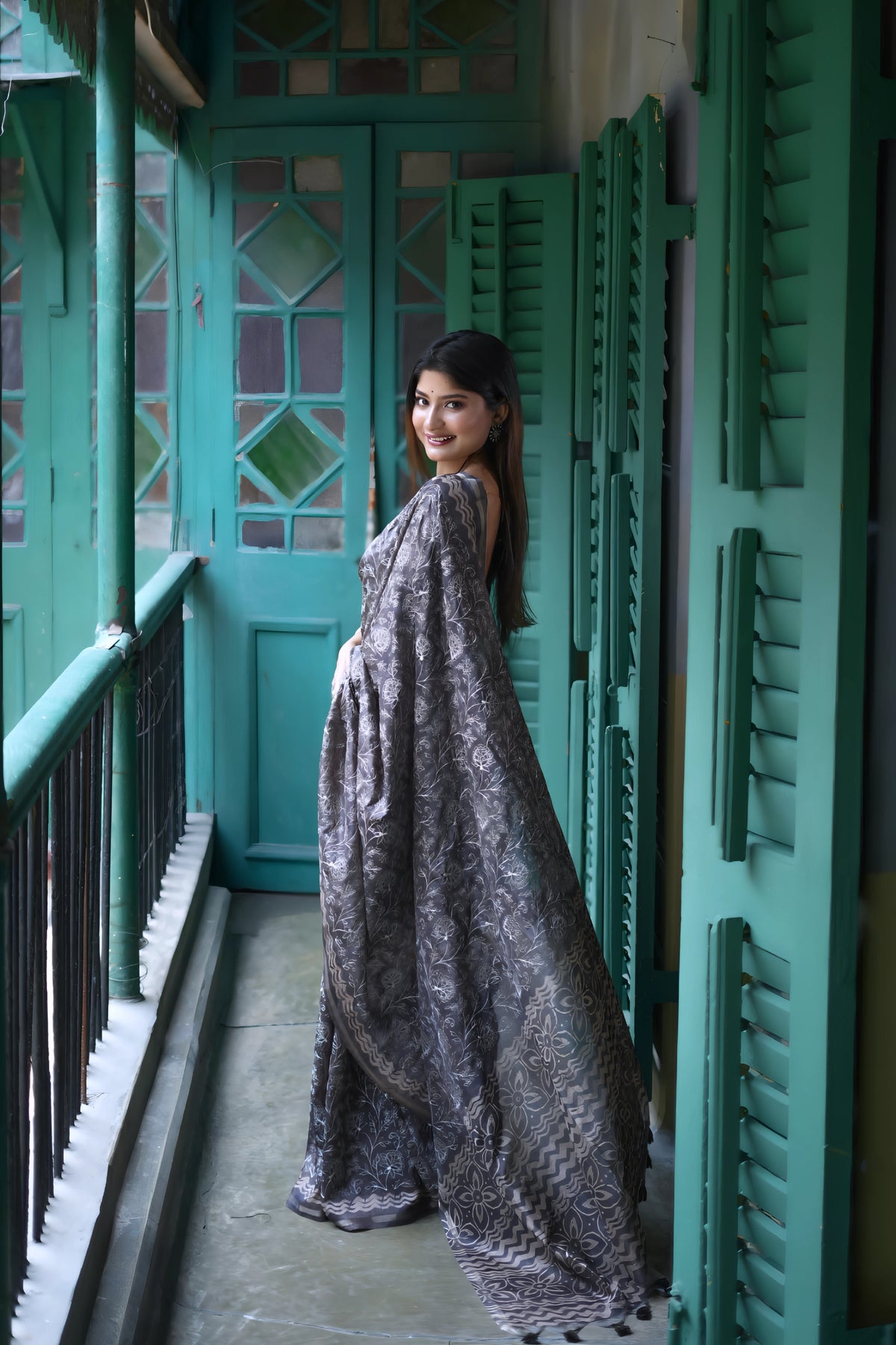 A woman models an elegant Black Tussar Silk Saree. The saree features dense all-over floral embroidery/weaving in a shimmering metallic silver thread. She wears a black sheer-sleeve blouse and heavy oxidized silver jewelry, including a large ring and stacked bangles, posing in a doorway framed by vibrant teal shutters.