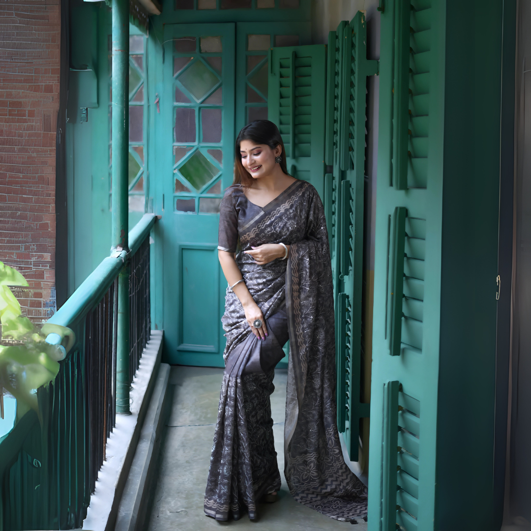 A woman models an elegant Black Tussar Silk Saree. The saree features dense all-over floral embroidery/weaving in a shimmering metallic silver thread. She wears a black sheer-sleeve blouse and heavy oxidized silver jewelry, including a large ring and stacked bangles, posing in a doorway framed by vibrant teal shutters.