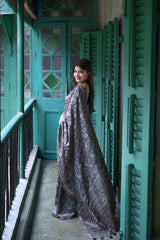 A woman models an elegant Black Tussar Silk Saree. The saree features dense all-over floral embroidery/weaving in a shimmering metallic silver thread. She wears a black sheer-sleeve blouse and heavy oxidized silver jewelry, including a large ring and stacked bangles, posing in a doorway framed by vibrant teal shutters.
