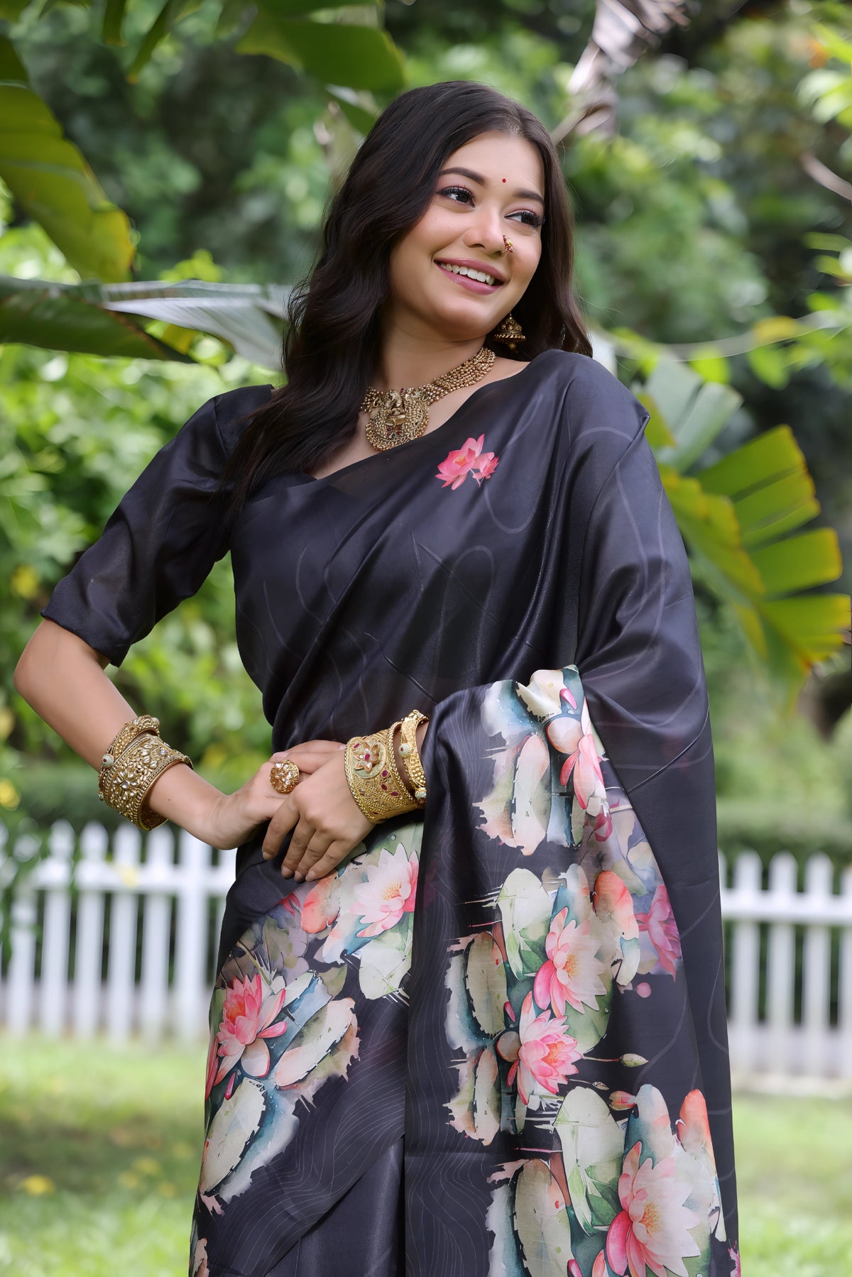 A woman models a stunning Deep Charcoal Black Soft Tussar Silk Saree. The saree features a large-scale digital print of colorful floral and lotus motifs in pink, coral, white, and green on the pallu and lower drape. She wears a matching elbow-sleeve black blouse and heavy gold Kundan jewelry, posing outdoors in a garden setting.