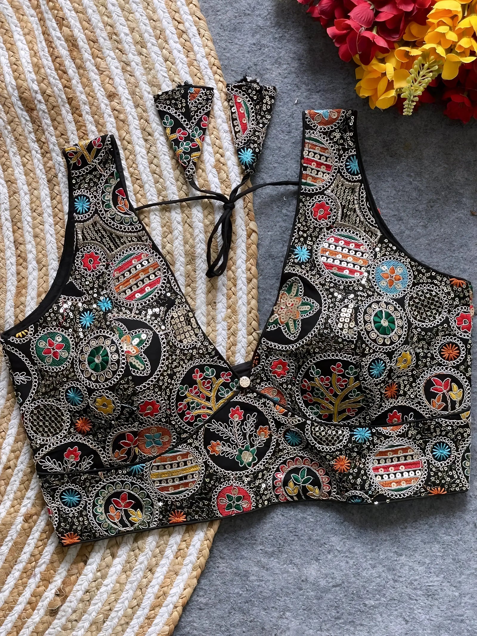 Traditional black blouse featuring vibrant Madhubani-inspired multicolor embroidery with circular floral and geometric motifs, deep V-neck, and tie-back detail, displayed flat on a textured mat with artificial red and yellow flowers in the corner.