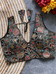 Traditional black blouse featuring vibrant Madhubani-inspired multicolor embroidery with circular floral and geometric motifs, deep V-neck, and tie-back detail, displayed flat on a textured mat with artificial red and yellow flowers in the corner.
