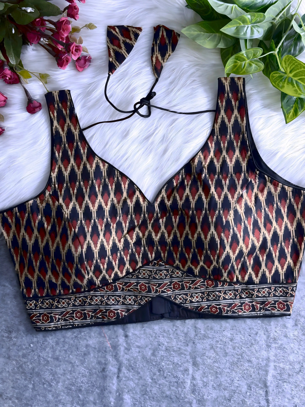 Traditional black and maroon silk blouse featuring Ikat-inspired geometric patterns in cream, gold, and red, wide shoulder straps, structured cups with a deep V-neckline, and a detailed patterned border at the hem. Includes a matching patterned tasseled tie-back, displayed flat on a soft white fur background with artificial pink and green foliage at the corners.