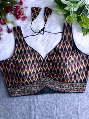 Traditional black and maroon silk blouse featuring Ikat-inspired geometric patterns in cream, gold, and red, wide shoulder straps, structured cups with a deep V-neckline, and a detailed patterned border at the hem. Includes a matching patterned tasseled tie-back, displayed flat on a soft white fur background with artificial pink and green foliage at the corners.