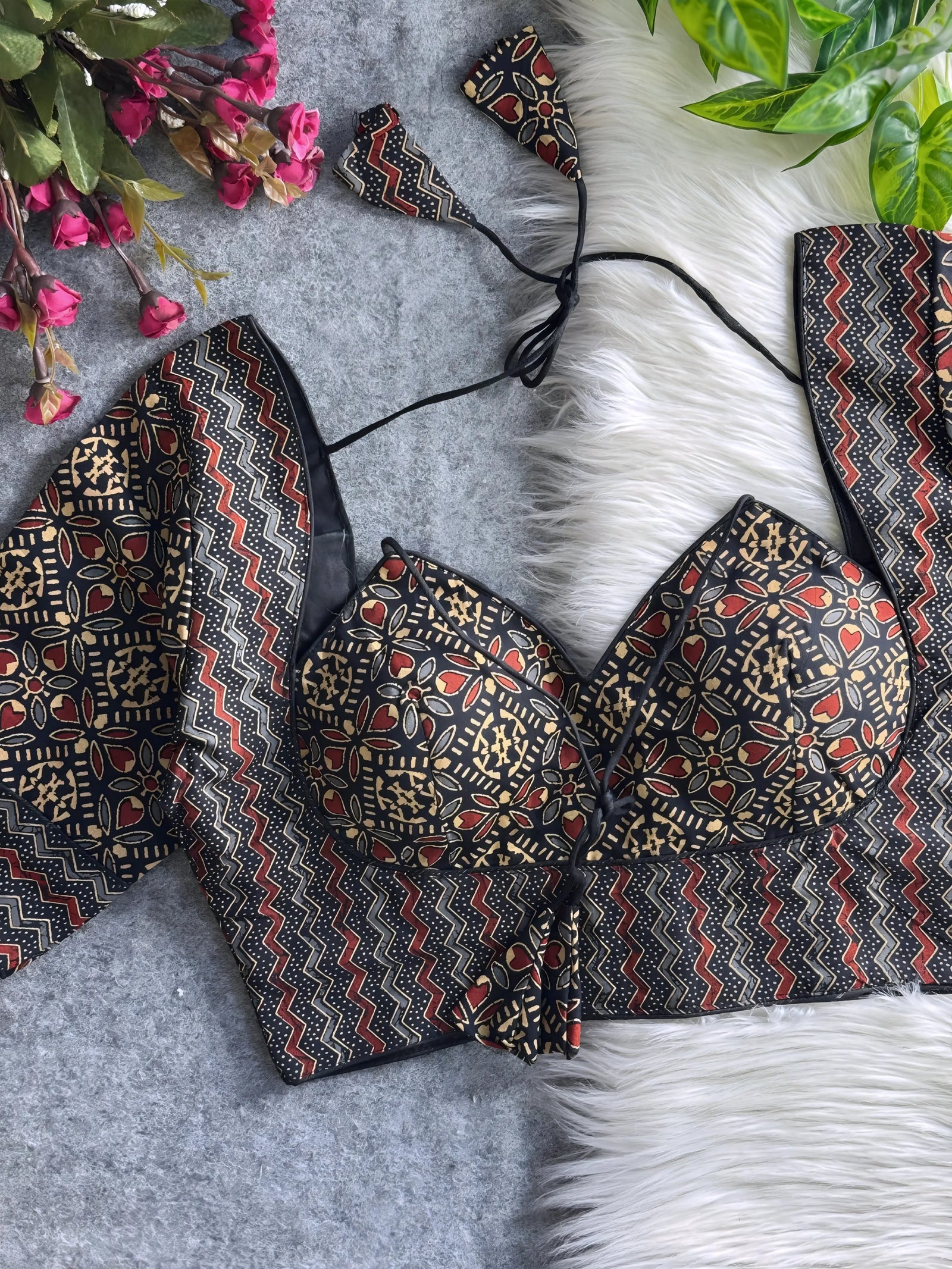 Black and maroon printed padded blouse with geometric and floral patterns, displayed on a white fur surface with artificial flowers and green leaves in the background.