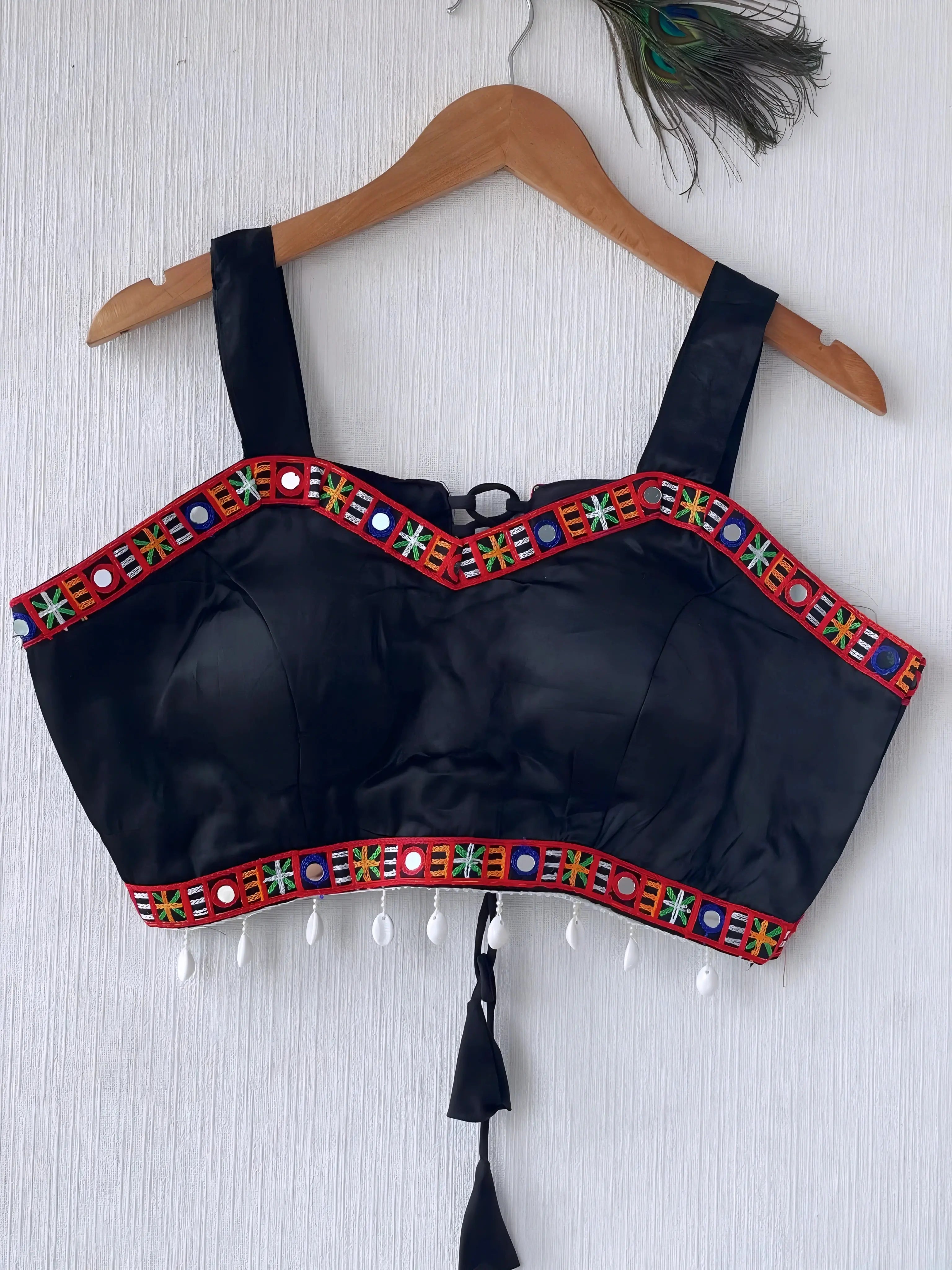 Black ethnic blouse featuring vibrant mirror work and multicolor embroidered border in red, green, blue, and yellow, adorned with pearl tassels and a tie-up at the back, displayed on a wooden hanger against a textured off-white wall with peacock feathers for a festive and traditional look.