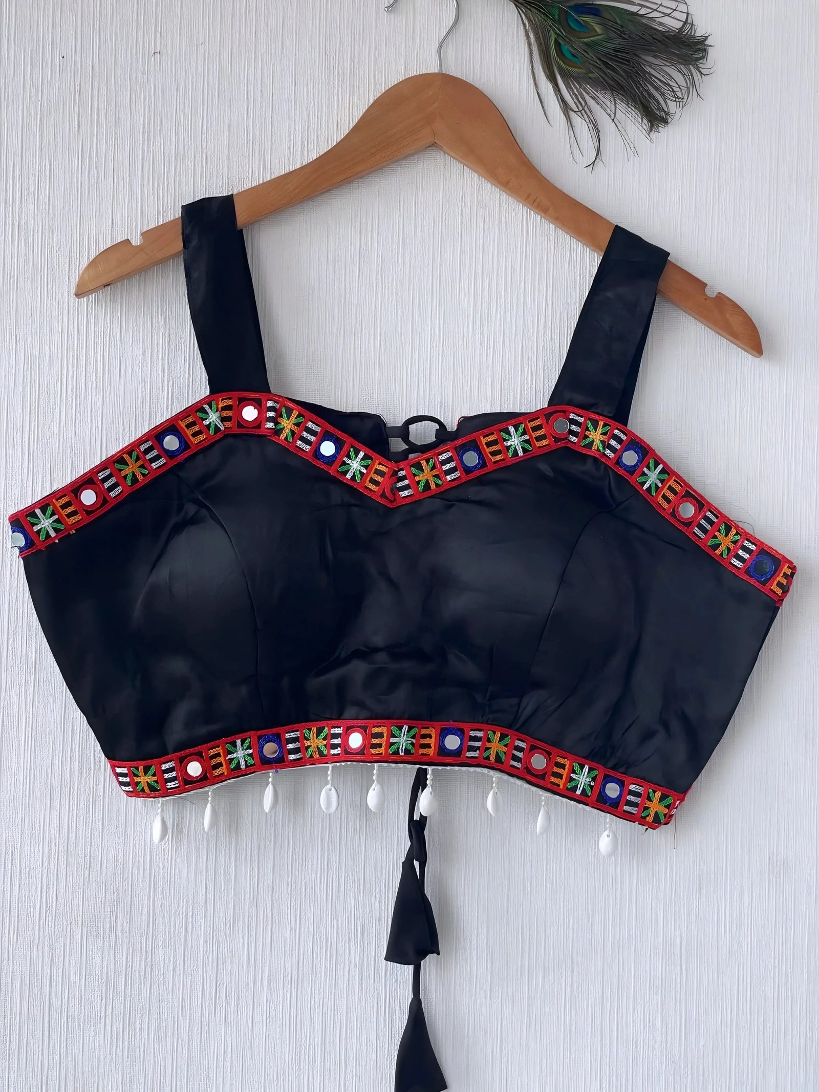 Black ethnic blouse featuring vibrant mirror work and multicolor embroidered border in red, green, blue, and yellow, adorned with pearl tassels and a tie-up at the back, displayed on a wooden hanger against a textured off-white wall with peacock feathers for a festive and traditional look.