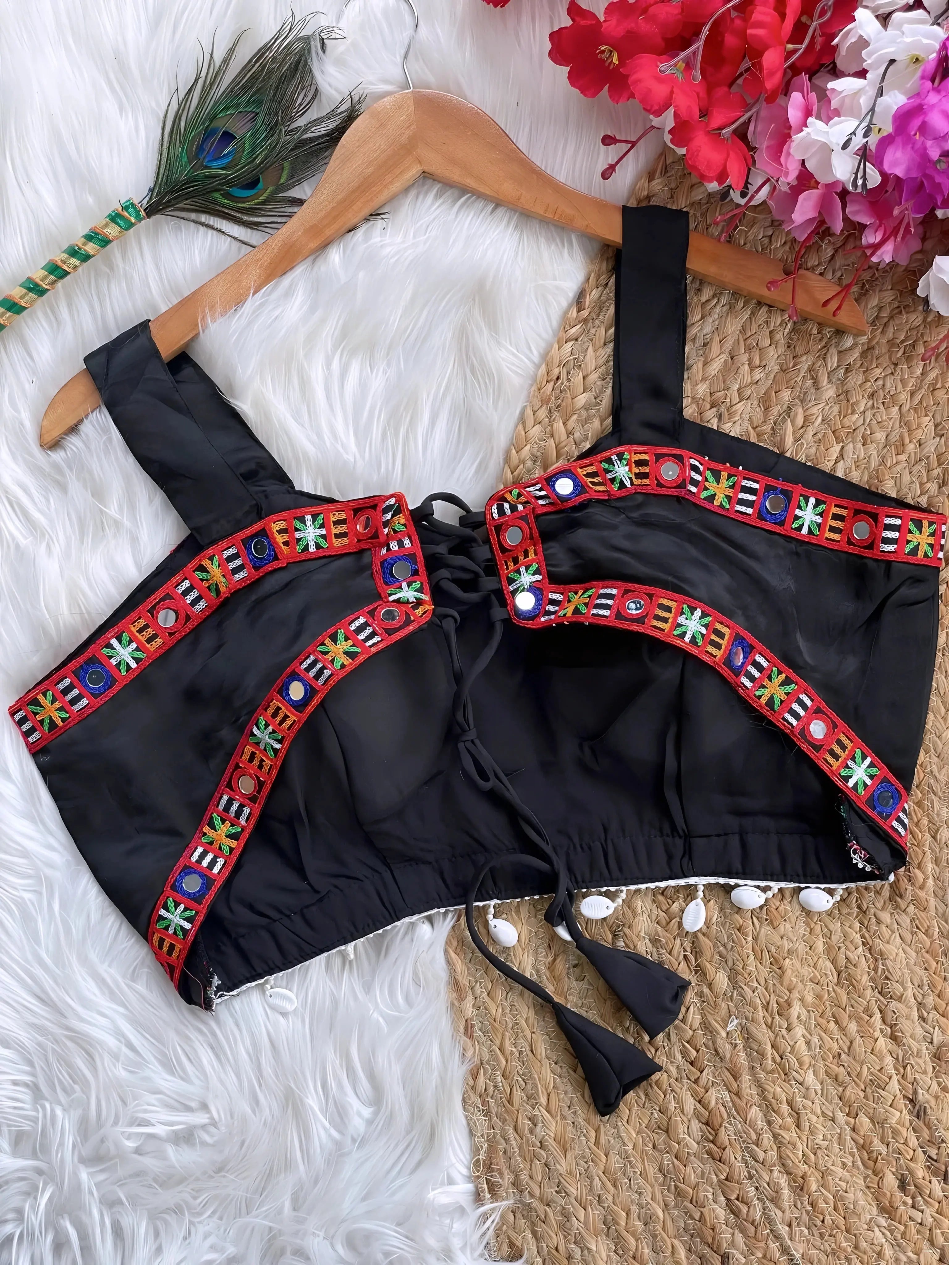 Black ethnic blouse featuring vibrant mirror work and multicolor embroidered border in red, green, blue, and yellow, adorned with pearl tassels and a tie-up at the back, displayed on a wooden hanger against a textured off-white wall with peacock feathers for a festive and traditional look.