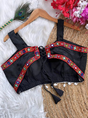 Black ethnic blouse featuring vibrant mirror work and multicolor embroidered border in red, green, blue, and yellow, adorned with pearl tassels and a tie-up at the back, displayed on a wooden hanger against a textured off-white wall with peacock feathers for a festive and traditional look.