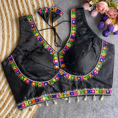 Black blouse featuring vibrant mirror work and traditional Kutch multicolor embroidery along the neckline and hem, adorned with scattered sequin details and shell tassels. The blouse includes a stylish back tie-up with matching embroidered tassels and is displayed flat on a textured surface with a floral bouquet, highlighting its festive ethnic appeal.