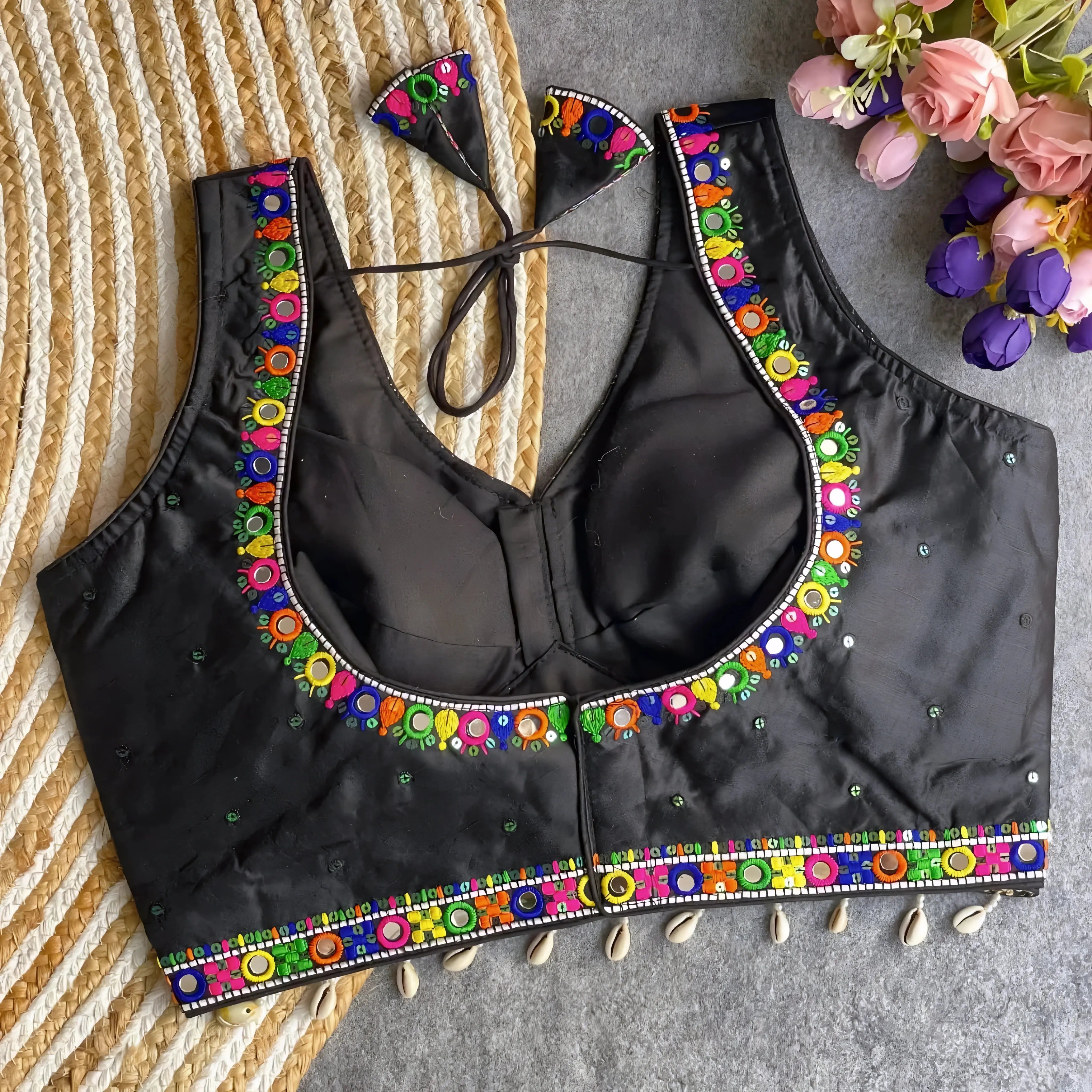 Black blouse featuring vibrant mirror work and traditional Kutch multicolor embroidery along the neckline and hem, adorned with scattered sequin details and shell tassels. The blouse includes a stylish back tie-up with matching embroidered tassels and is displayed flat on a textured surface with a floral bouquet, highlighting its festive ethnic appeal.