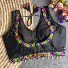 Black blouse featuring vibrant mirror work and traditional Kutch multicolor embroidery along the neckline and hem, adorned with scattered sequin details and shell tassels. The blouse includes a stylish back tie-up with matching embroidered tassels and is displayed flat on a textured surface with a floral bouquet, highlighting its festive ethnic appeal.