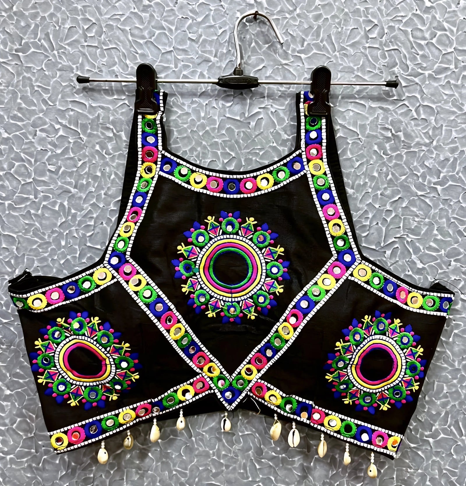Black blouse featuring vibrant circular and oval mirror work with traditional Kutch multicolor embroidery, adorned with bold geometric borders and shell tassel drops along the hem. Displayed on a hanger against a textured silver background, highlighting its festive and culturally inspired ethnic design.