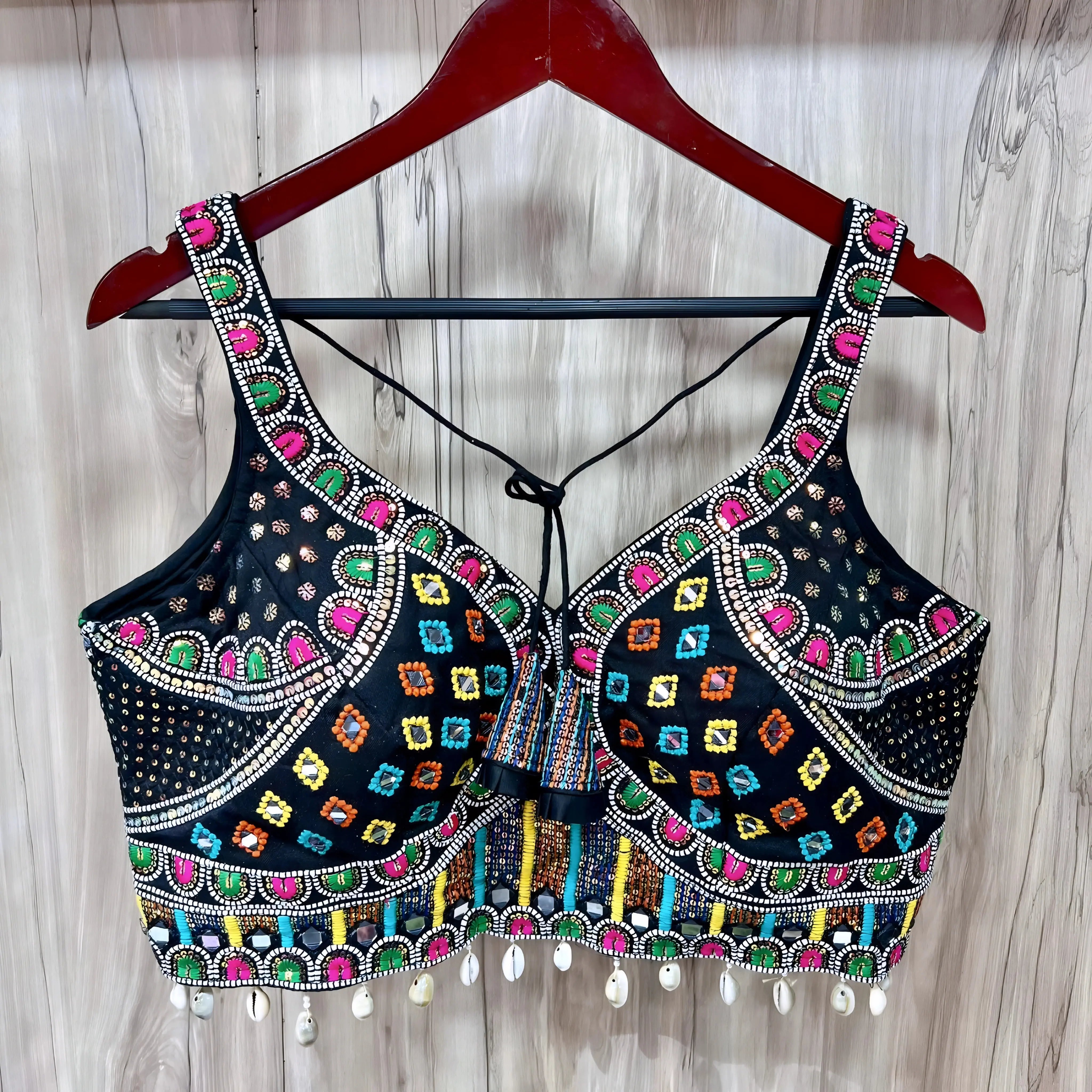 Traditional black blouse featuring vibrant multicolor Kutch-style embroidery with geometric patterns in yellow, orange, blue, green, and pink, accented with intricate beadwork, white cowrie shell trim along the hem, and decorative tie-back detail. The blouse is displayed on a wooden hanger against a light wood background.