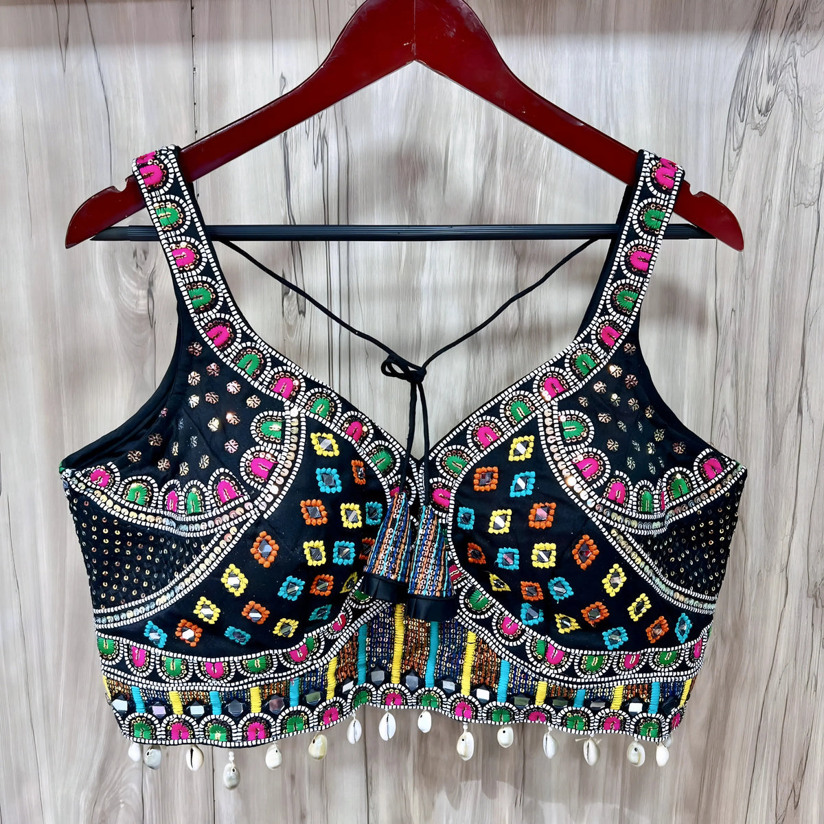 Traditional black blouse featuring vibrant multicolor Kutch-style embroidery with geometric patterns in yellow, orange, blue, green, and pink, accented with intricate beadwork, white cowrie shell trim along the hem, and decorative tie-back detail. The blouse is displayed on a wooden hanger against a light wood background.