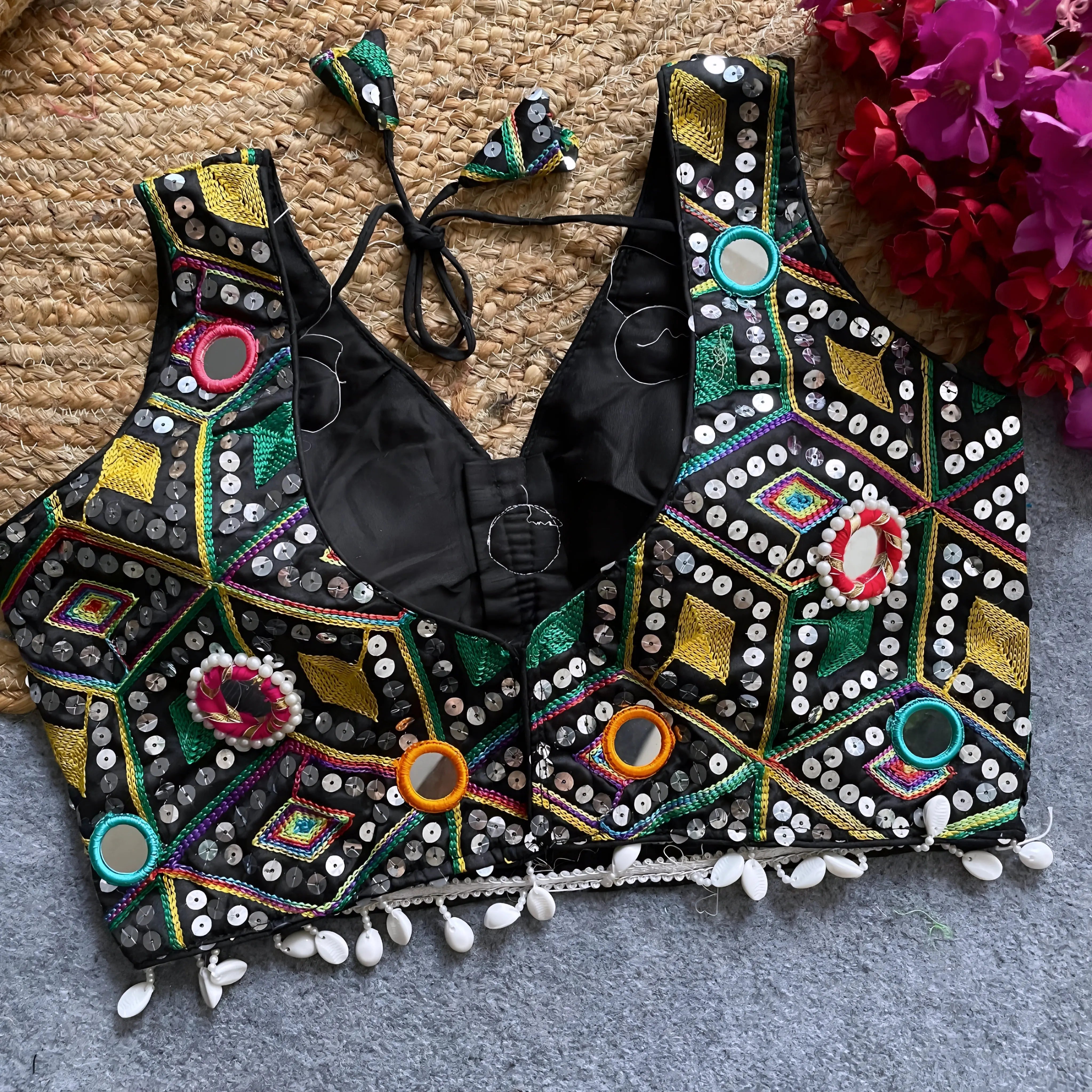 Traditional black Gujarati-style blouse richly adorned with vibrant multicolor geometric embroidery and circular mirror work, decorated with colorful thread rings, white cowrie shell trim along the hem, and tasseled tie-back detail, displayed flat on a grey carpet with a woven mat and fuchsia artificial flowers beside it.