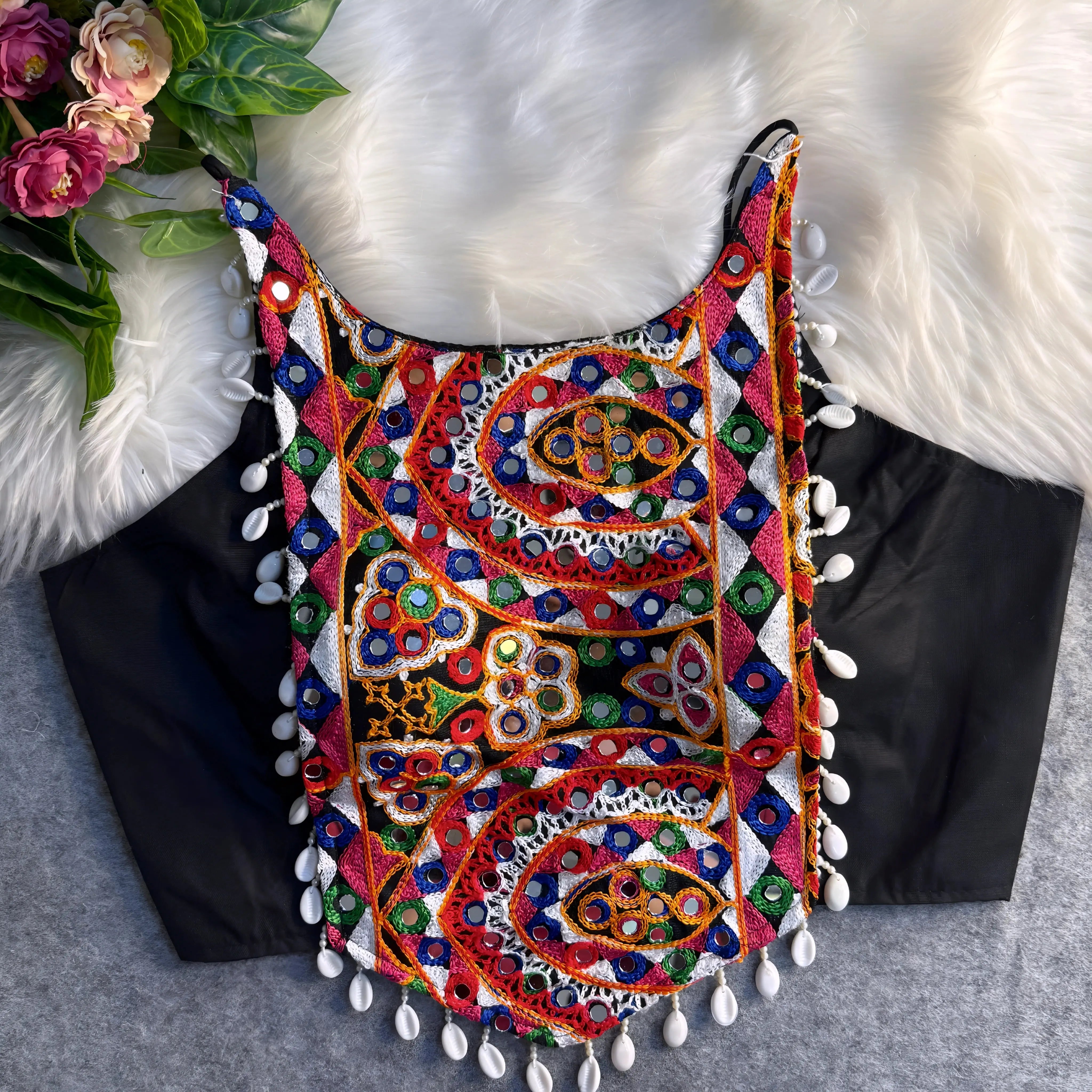Traditional black blouse featuring vibrant multicolor mirror work and intricate thread embroidery in geometric and paisley patterns, accented with white shell tassels along the hem, displayed flat on a soft fur and grey background with floral and leafy decor.