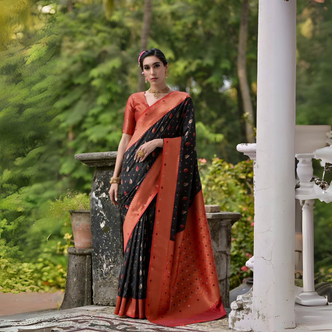 A woman models a striking Black Banarasi Silk Saree with scattered red and gold Zari motifs. The saree is framed by a wide, opulent Terracotta Red border and pallu featuring dense traditional gold weaving. She wears a red blouse and heavy gold Kundan jewelry, posing outdoors on a balcony.