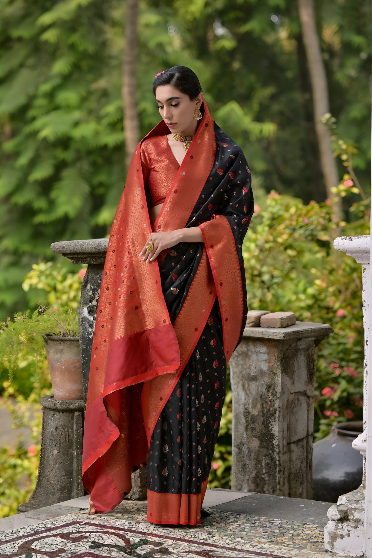 A woman models a striking Black Banarasi Silk Saree with scattered red and gold Zari motifs. The saree is framed by a wide, opulent Terracotta Red border and pallu featuring dense traditional gold weaving. She wears a red blouse and heavy gold Kundan jewelry, posing outdoors on a balcony.