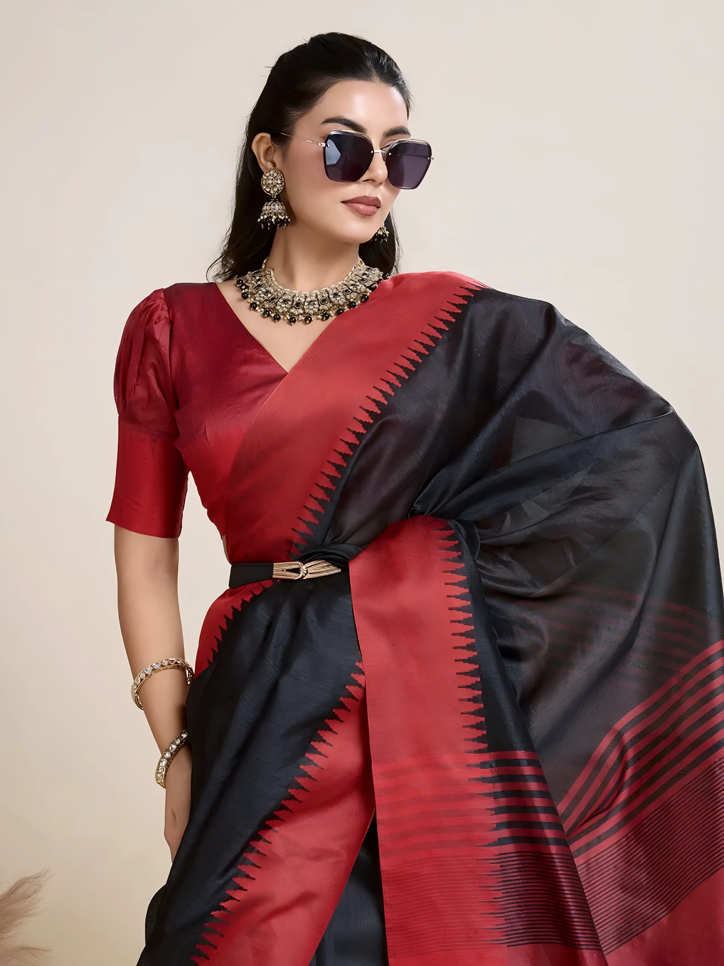 A woman models a striking Black Raw Silk Saree featuring a wide, vibrant deep red border with traditional temple weaving. The saree is cinched with a belt. She wears a red blouse, a metallic choker necklace, and large sunglasses, posing indoors.