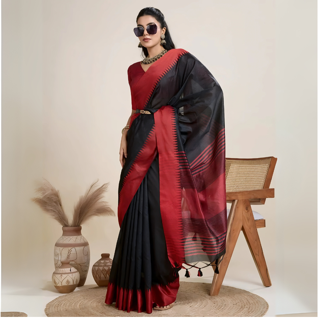 A woman models a striking Black Raw Silk Saree featuring a wide, vibrant deep red border with traditional temple weaving. The saree is cinched with a belt. She wears a red blouse, a metallic choker necklace, and large sunglasses, posing indoors.