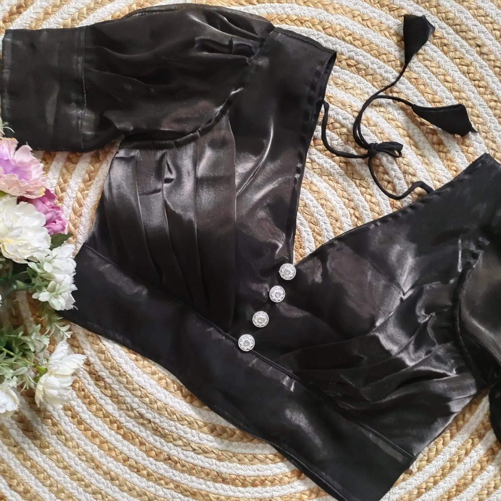 Black satin blouse with pleated detailing and three decorative silver buttons on the front, featuring elbow-length sleeves and tie-up tassels, displayed on a textured circular mat with pink and white flowers on the side.