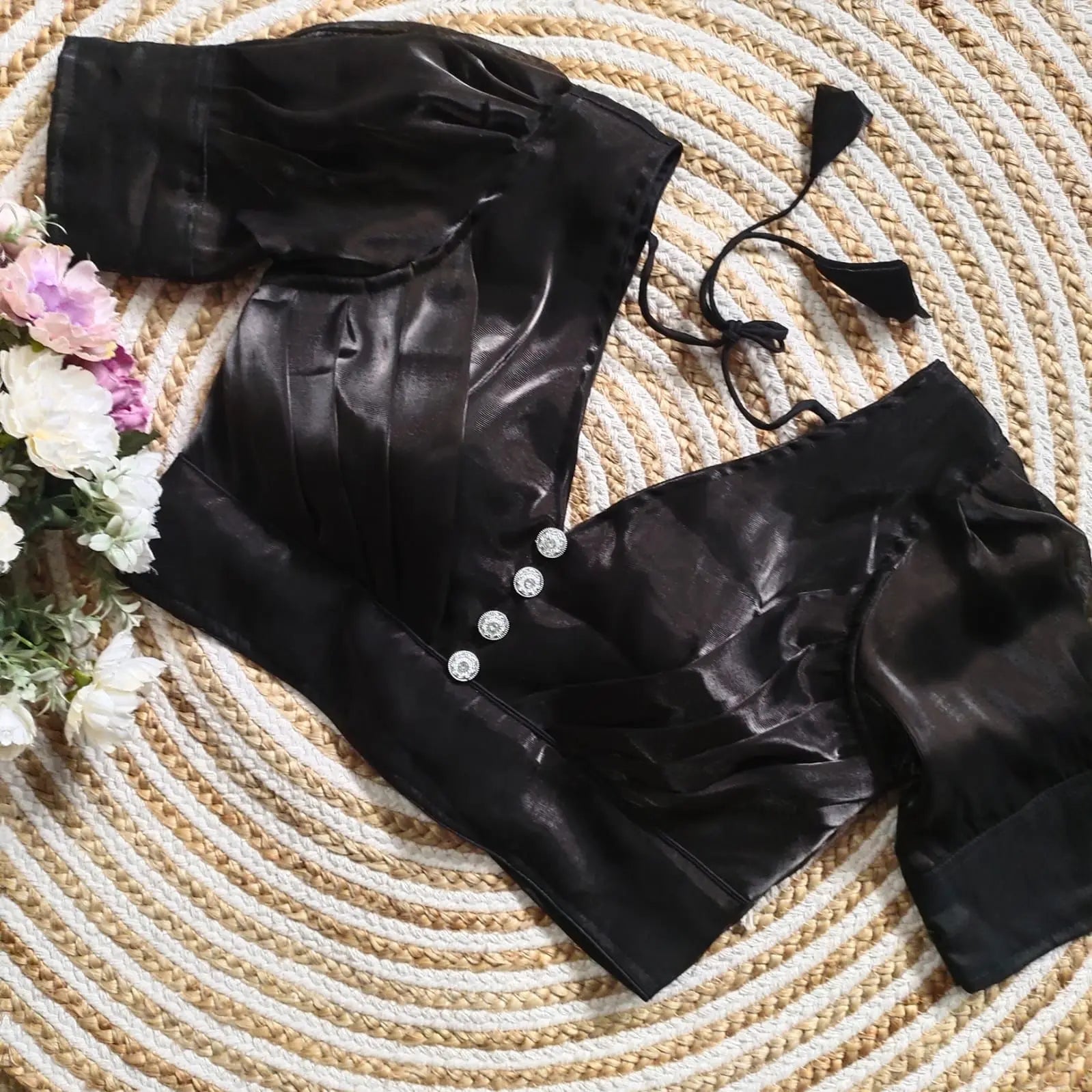 Black satin blouse with pleated detailing and three decorative silver buttons on the front, featuring elbow-length sleeves and tie-up tassels, displayed on a textured circular mat with pink and white flowers on the side.