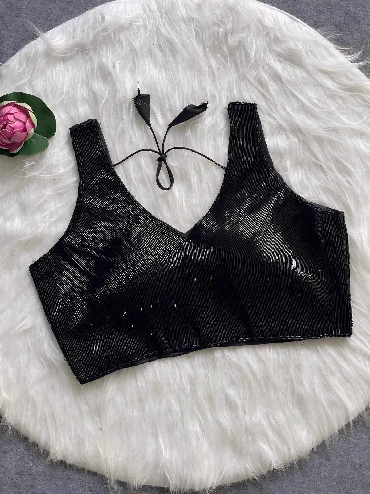  Black sequin sleeveless blouse with tie-back detail, displayed on a white fur background with a pink floral prop in the corner.