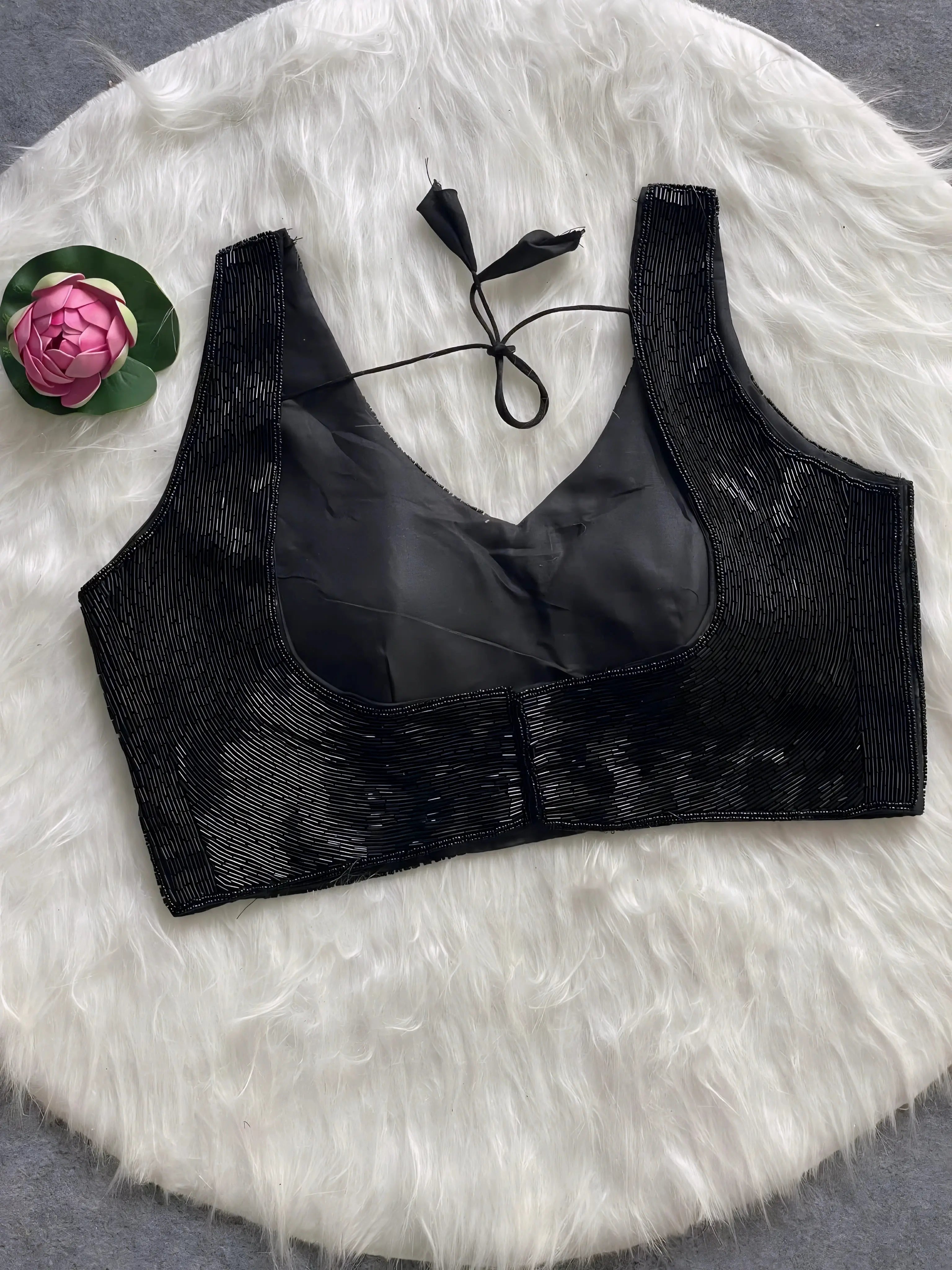  Black sequin sleeveless blouse with tie-back detail, displayed on a white fur background with a pink floral prop in the corner