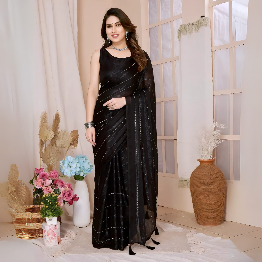 A woman models a chic, sheer Black Rimzim Silk Saree. The saree body features subtle vertical stripes and scattered embellishments of fine metallic threadwork (Zari/Lurex). She wears a matching black sleeveless blouse and accessorizes with chunky oxidized silver jewelry, including a layered necklace and stacked bangles, posing indoors.