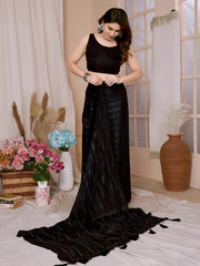 A woman models a chic, sheer Black Rimzim Silk Saree. The saree body features subtle vertical stripes and scattered embellishments of fine metallic threadwork (Zari/Lurex). She wears a matching black sleeveless blouse and accessorizes with chunky oxidized silver jewelry, including a layered necklace and stacked bangles, posing indoors.