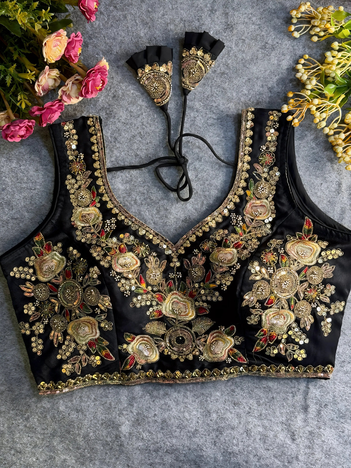 Traditional black silk blouse featuring intricate gold zardozi embroidery and beadwork with multicolor floral and leaf motifs on the front and neckline, finished with decorative tassel ties, displayed flat on a textured grey background with artificial pink and yellow flowers in the corners.