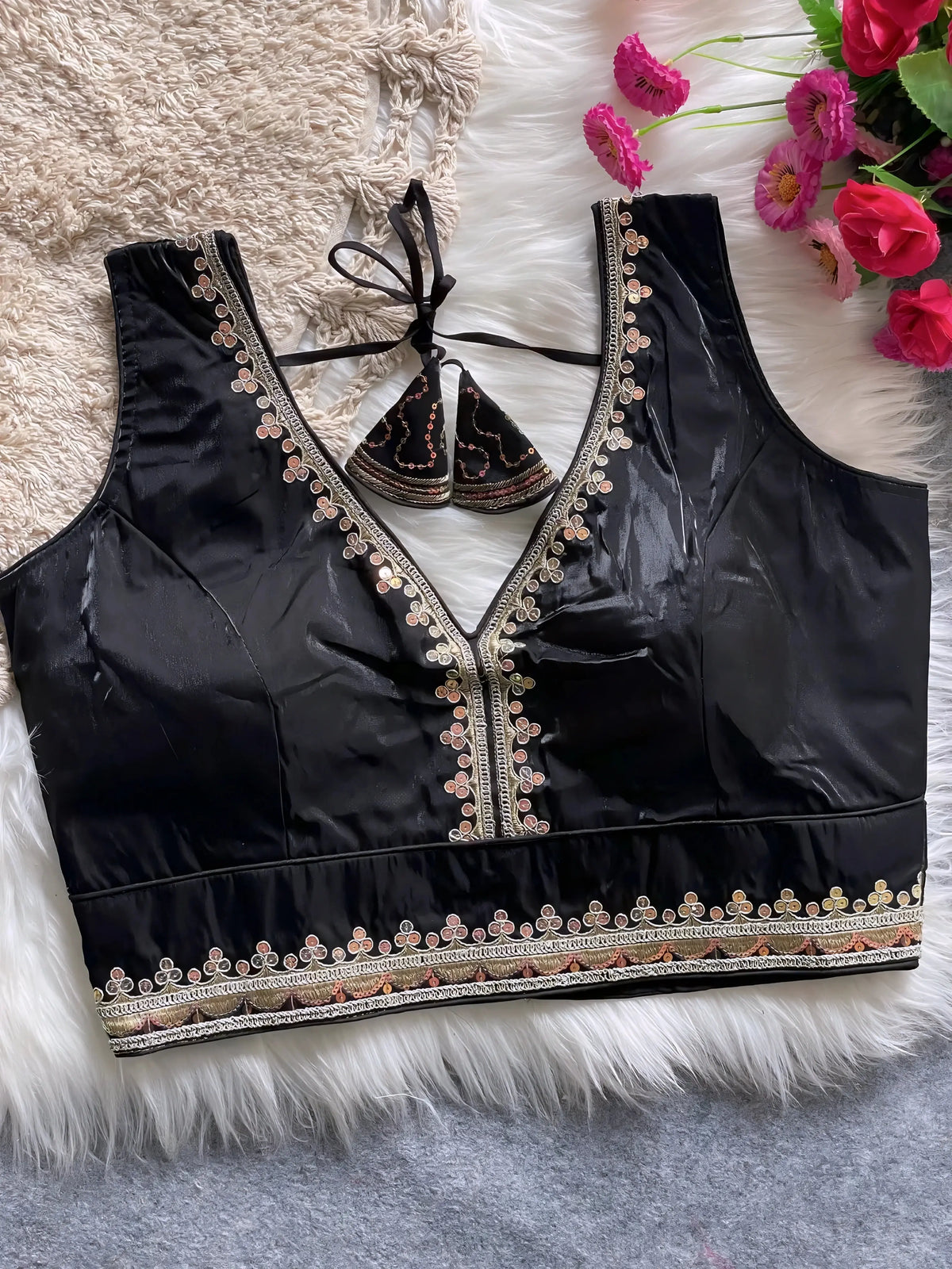 Traditional black silk blouse featuring intricate multicolor mirror work and thread embroidery along the neckline and hem, with decorative tassel tie at the back, displayed flat on a soft fur and textured mat background with artificial pink flowers in the corner.