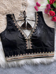 Traditional black silk blouse featuring intricate multicolor mirror work and thread embroidery along the neckline and hem, with decorative tassel tie at the back, displayed flat on a soft fur and textured mat background with artificial pink flowers in the corner.
