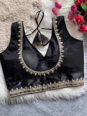 Traditional black silk blouse featuring intricate multicolor mirror work and thread embroidery along the neckline and hem, with decorative tassel tie at the back, displayed flat on a soft fur and textured mat background with artificial pink flowers in the corner.
