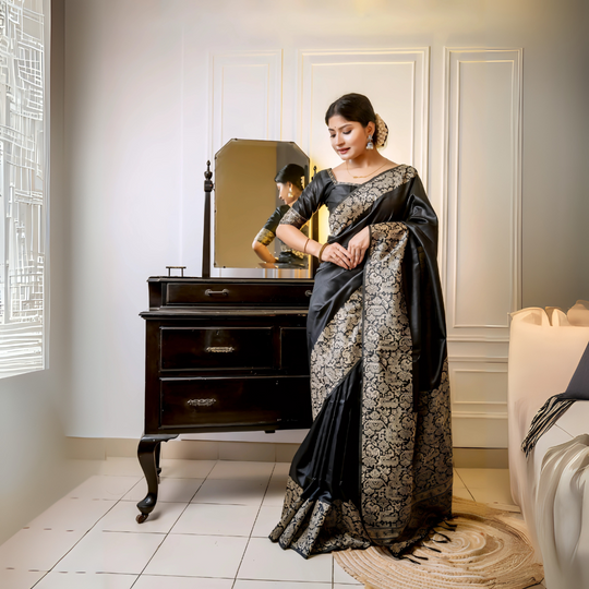A woman models a luxurious Deep Black Raw Silk Saree. The saree features a wide border and pallu with dense, intricate gold Zari weaving in a Kalamkari-style floral and paisley pattern. She wears a matching short-sleeve black blouse and elegant gold jewelry, with white flowers adorning her hair, posing indoors next to a dark wooden vanity.