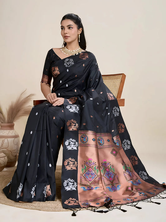 A woman models a luxurious Black Soft Silk Saree. The saree features dual-toned copper and silver metallic woven motifs on the body, and a wide, contrasting peach/rose gold pallu panel richly woven with multi-colored traditional designs. She wears a matching black blouse and heavy gold Kundan jewelry.
