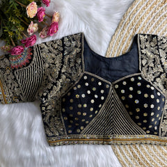 Traditional black silk blouse featuring intricate gold zardosi embroidery with floral motifs, sequin polka dot patterns, geometric pinstripes, and a colorful peacock feather motif on the sleeve, displayed flat on a white fur surface with a braided mat and artificial pink flowers in the corner.