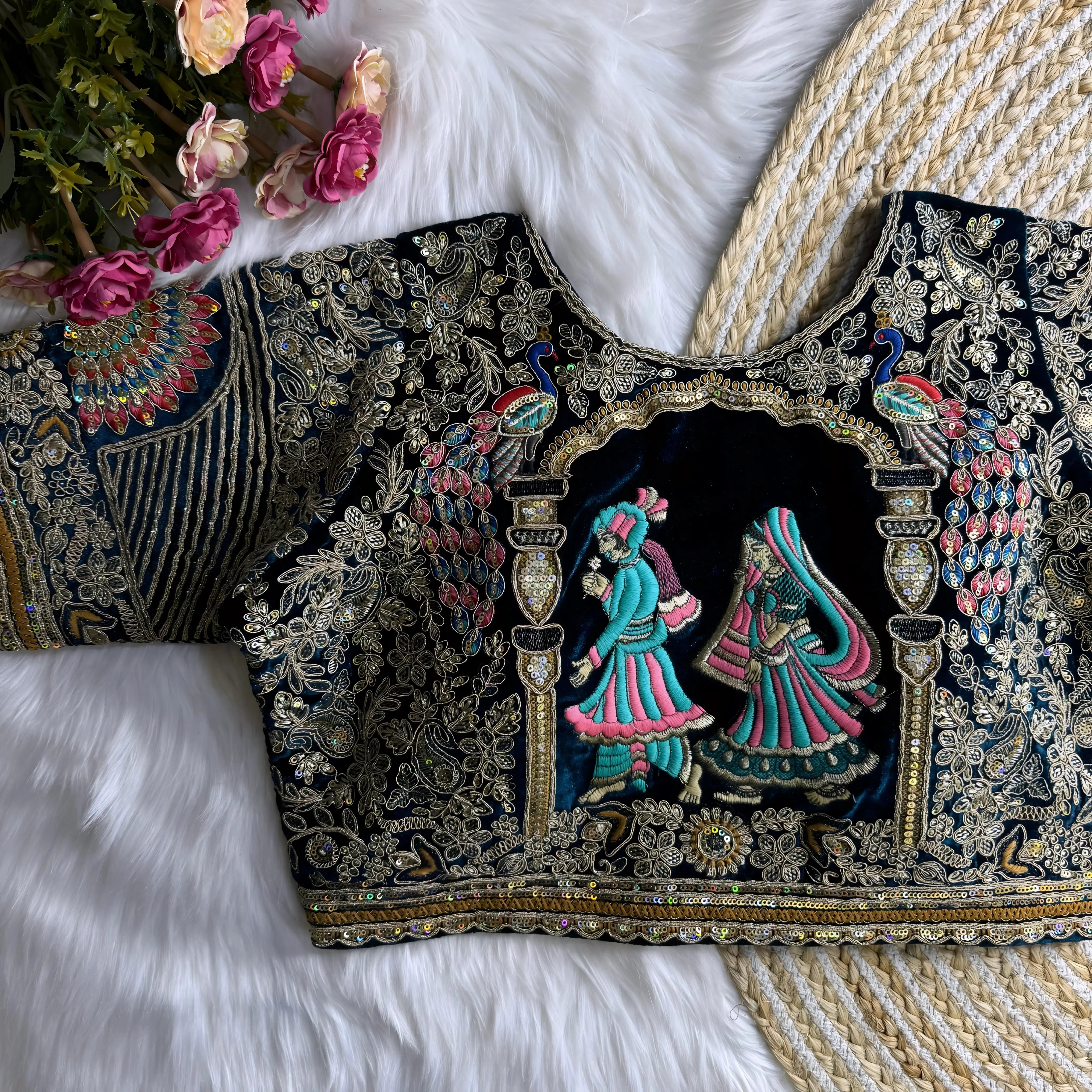 Traditional black silk blouse featuring intricate gold zardosi embroidery with floral motifs, sequin polka dot patterns, geometric pinstripes, and a colorful peacock feather motif on the sleeve, displayed flat on a white fur surface with a braided mat and artificial pink flowers in the corner.