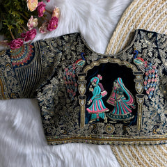 Traditional black silk blouse featuring intricate gold zardosi embroidery with floral motifs, sequin polka dot patterns, geometric pinstripes, and a colorful peacock feather motif on the sleeve, displayed flat on a white fur surface with a braided mat and artificial pink flowers in the corner.
