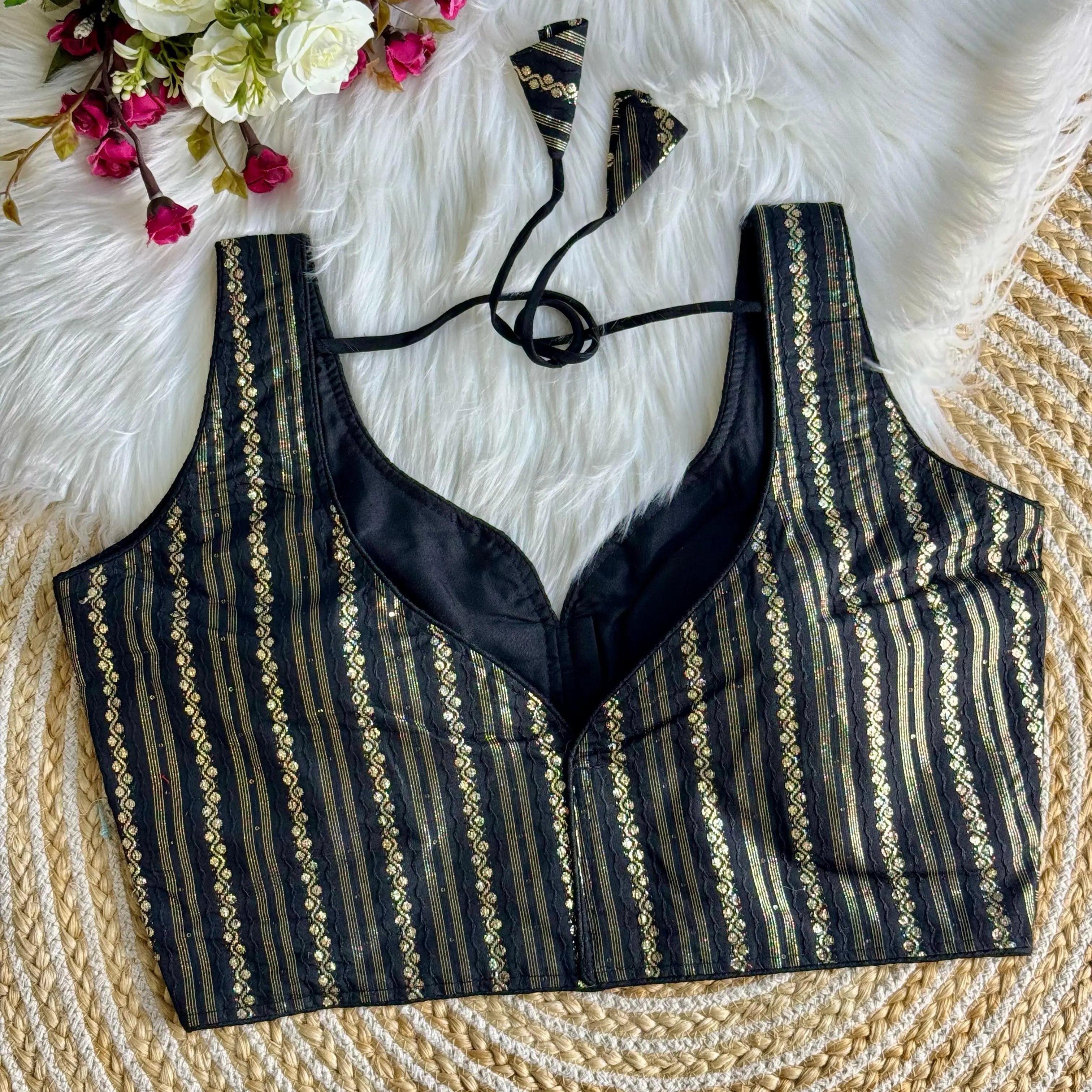 Black blouse top with intricate zari work design, featuring vertical stripes and floral patterns, displayed on a white fur background with a bouquet of white and pink flowers.