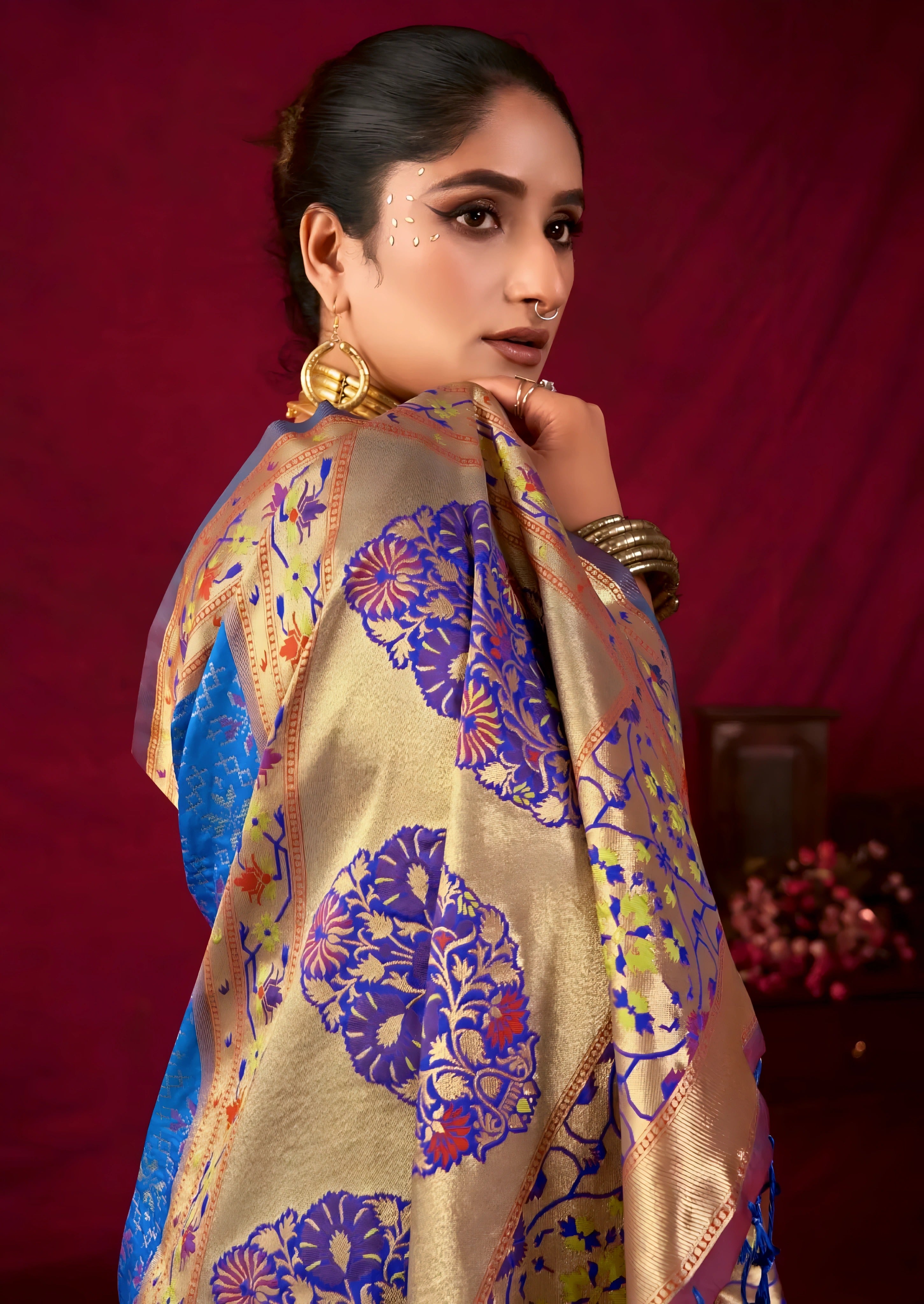 A woman models a luxurious Royal Blue Soft Banarasi Silk Saree. The wide border and pallu feature dense gold Zari weaving with prominent purple accents and large traditional motifs. She wears a unique dual-toned (purple/mauve) blouse and a heavy gold Kundan choker necklace, posing indoors.