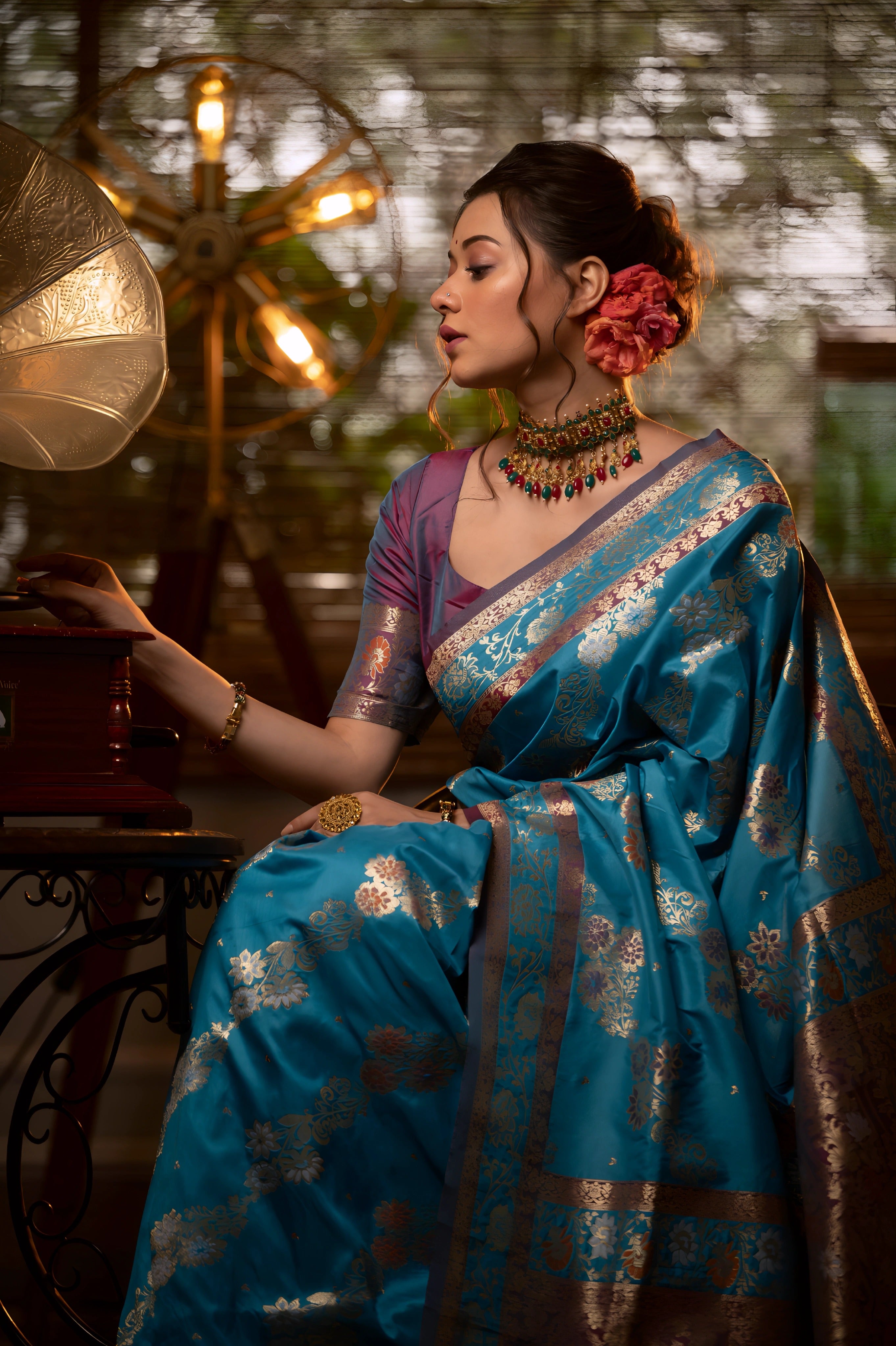 A woman models a luxurious Aqua Blue Soft Banarasi Silk Saree. The saree features heavy dual-toned gold and dark Zari weaving with a prominent dark brown/maroon accent border. She wears a contrasting mauve short-sleeve blouse and a heavy gold and stone choker necklace, posing in a dramatically lit vintage setting
