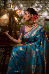 A woman models a luxurious Aqua Blue Soft Banarasi Silk Saree. The saree features heavy dual-toned gold and dark Zari weaving with a prominent dark brown/maroon accent border. She wears a contrasting mauve short-sleeve blouse and a heavy gold and stone choker necklace, posing in a dramatically lit vintage setting
