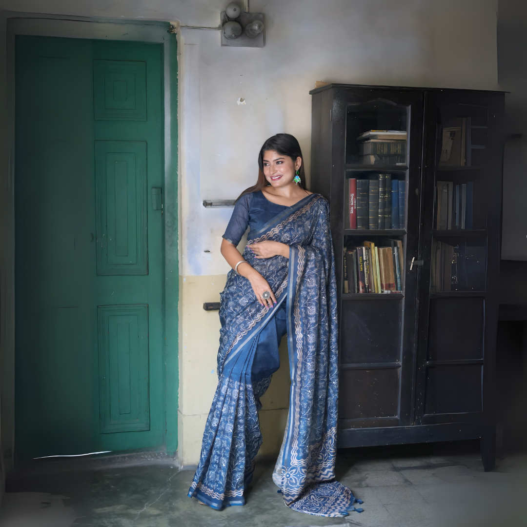 A woman models an elegant Navy Blue Tussar Silk Saree. The saree features dense all-over floral embroidery/weaving in a light metallic color, framed by a contrasting light metallic border. She wears a matching sheer-sleeve blue blouse and large oxidized silver earrings with blue accents, posing indoors next to a teal door and a dark wooden bookshelf.
