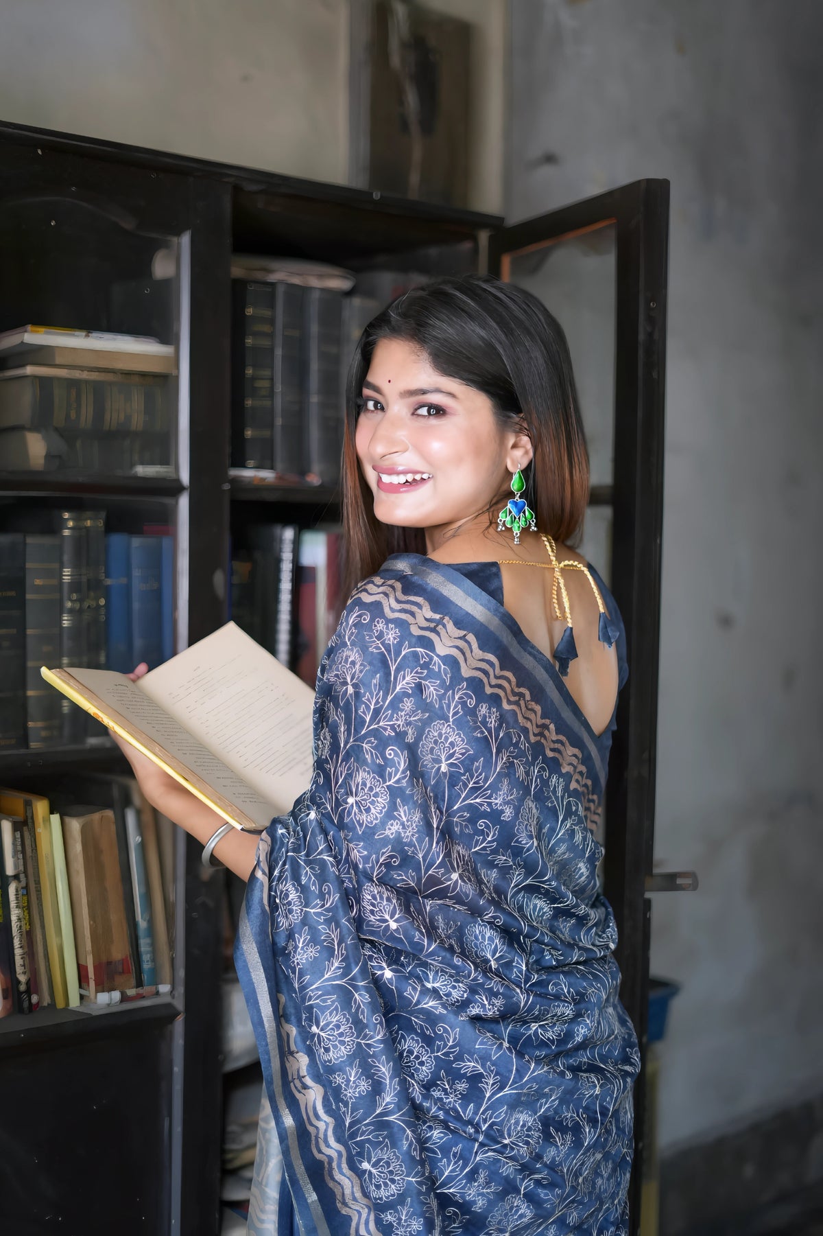 A woman models an elegant Navy Blue Tussar Silk Saree. The saree features dense all-over floral embroidery/weaving in a light metallic color, framed by a contrasting light metallic border. She wears a matching sheer-sleeve blue blouse and large oxidized silver earrings with blue accents, posing indoors next to a teal door and a dark wooden bookshelf.