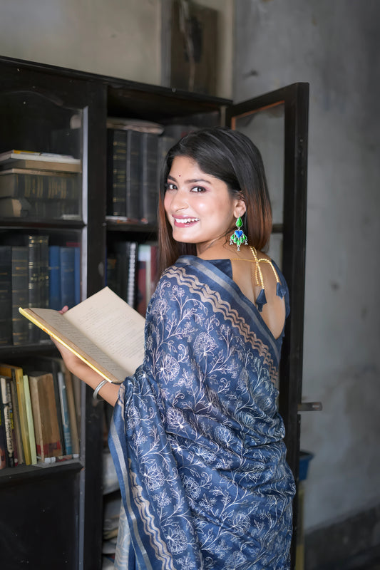 A woman models an elegant Navy Blue Tussar Silk Saree. The saree features dense all-over floral embroidery/weaving in a light metallic color, framed by a contrasting light metallic border. She wears a matching sheer-sleeve blue blouse and large oxidized silver earrings with blue accents, posing indoors next to a teal door and a dark wooden bookshelf.